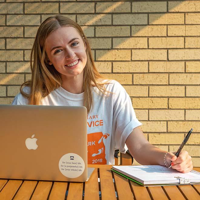 Smiling student outside with laptop and notebook on table in front of her