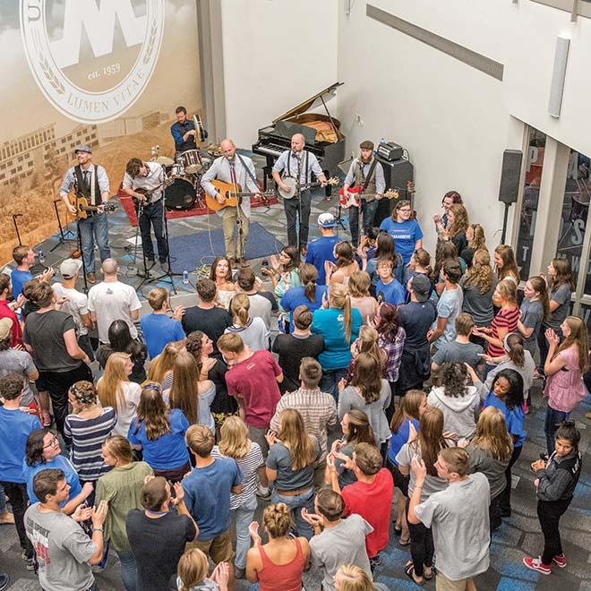 A band on stage playing for a large group of energetic students in the lower level of the university center