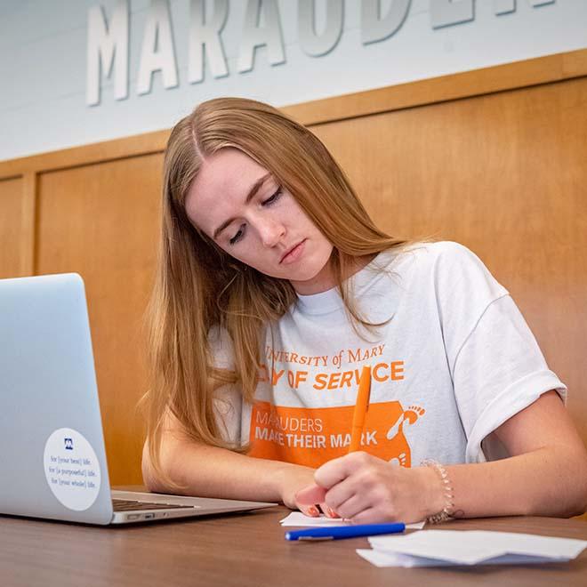 Female student writing notes with laptop on table in front of her.