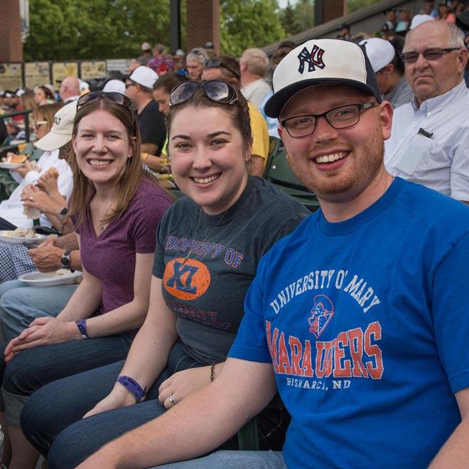 Three alumni smiling at a baseball game.