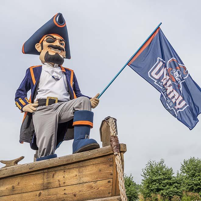 Max the Marauder standing at the front of a boat with a flag in hand.