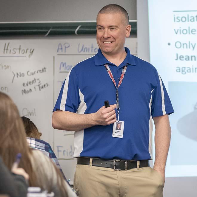 High school teacher smiling in front of classroom with presentation behind him