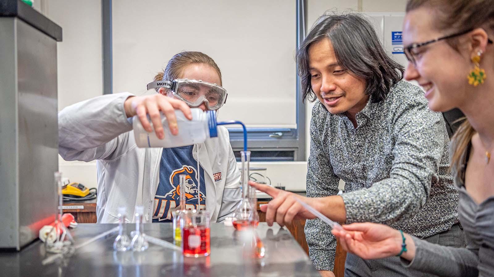 Chemistry student adding liquid to a beaker in a chemistry lab with professor and student watching