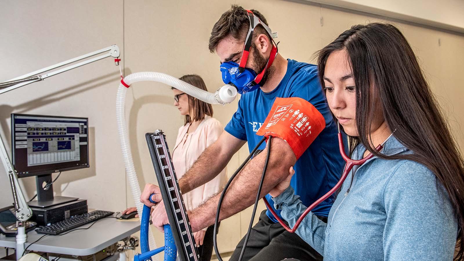 exercise science student monitoring the breathing of an athlete riding an exercise bike and wearing a VO2 max mask.