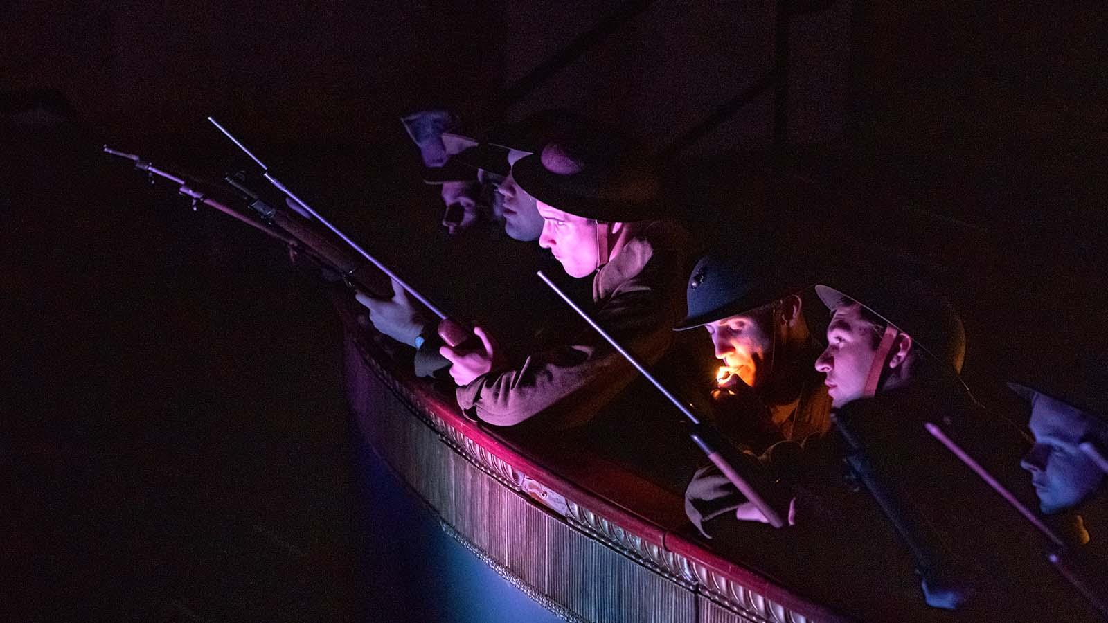 A closeup of eight actors in a WWI production leaning over a ledge with guns up and looking in the distance.