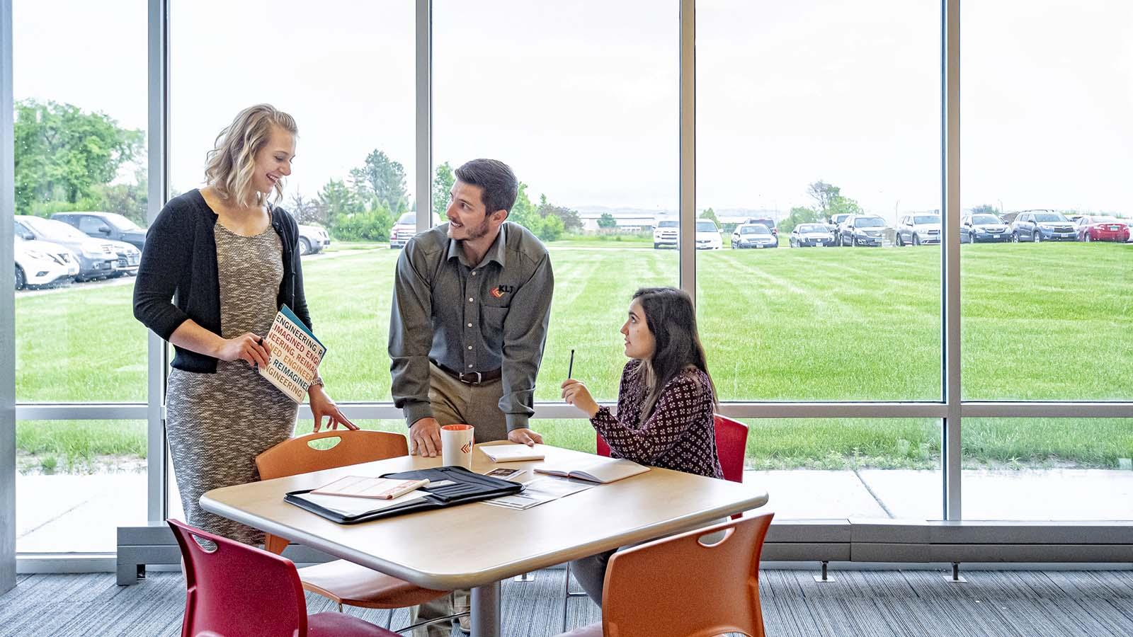 Three young employees meeting at a table with large windows behind them.