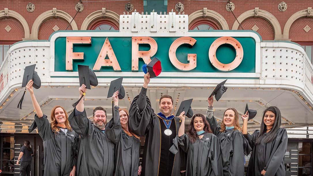 Fargo adult students in their graduation gowns standing and waving their graduation caps outside the Fargo Theater sign with University of Mary President Monsignor Shea
