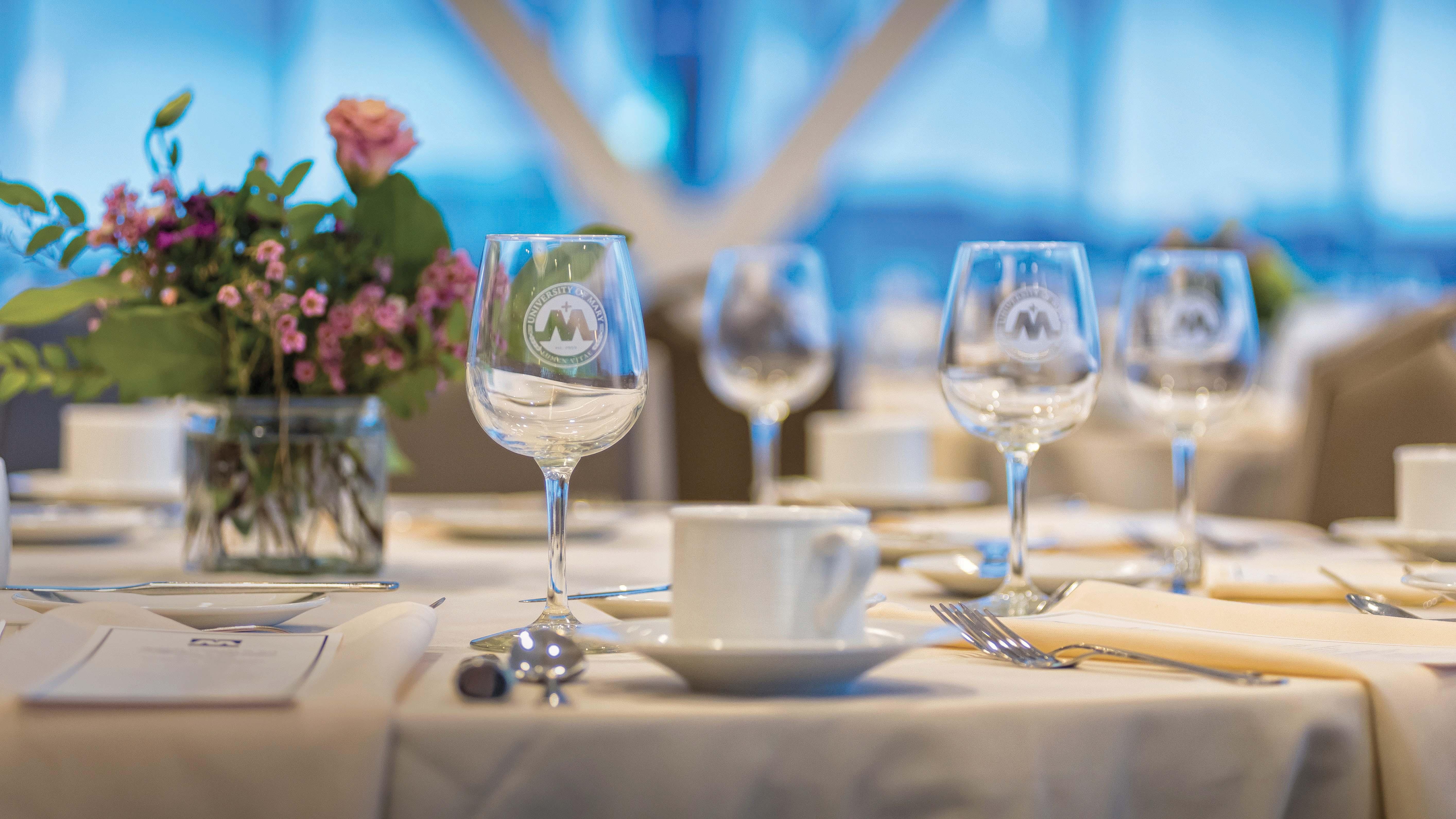 University of Mary wine glasses etched with its seal are set on a table with a centerpiece, menus, silverware, and napkins adding elegance to the formal dining decor inside Founders Hall on campus.