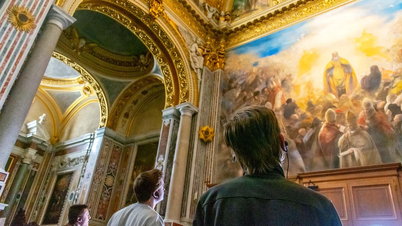 Three students studying abroad in Rome listening to commentary on headsets while admiring a classical painting in a museum