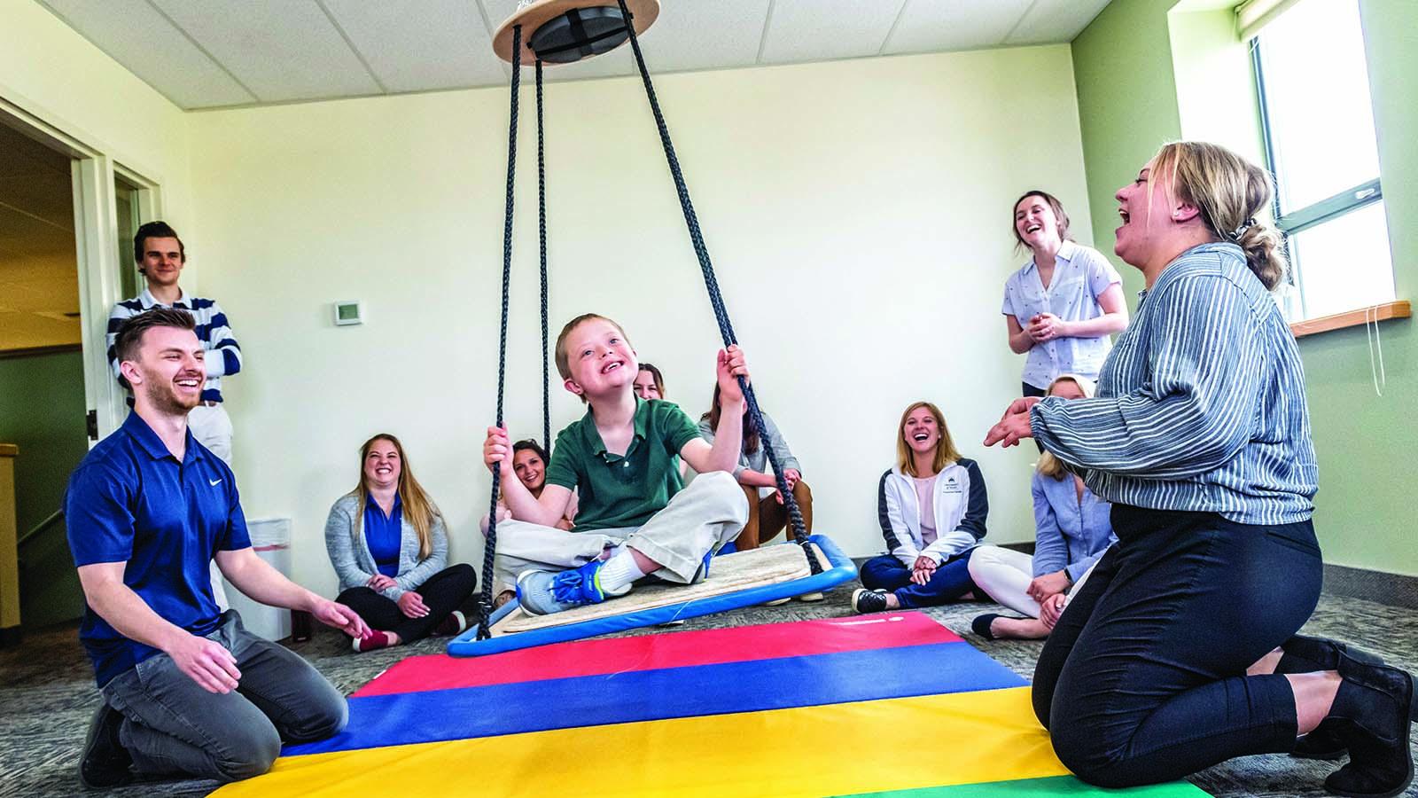 Several smiling occupational therapy students meeting with a child client who is swinging above a multi-colored mat