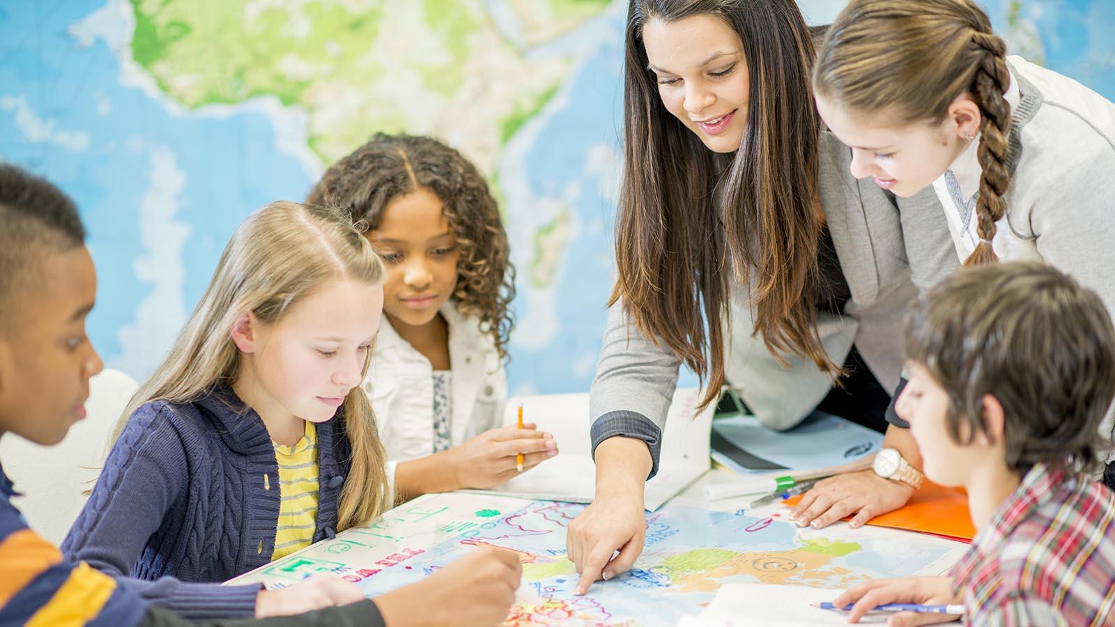 Elementary age children and their teacher sitting at a desk and drawing on a world map for history class.