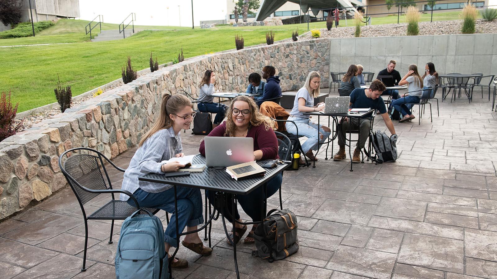 Several year-round campus students sitting at tables on a patio studying with laptops open