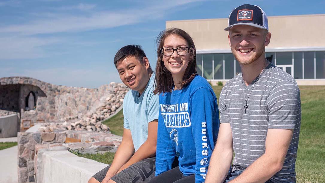 Three smiling students sitting on the edge of the rock wall leading to the grotto