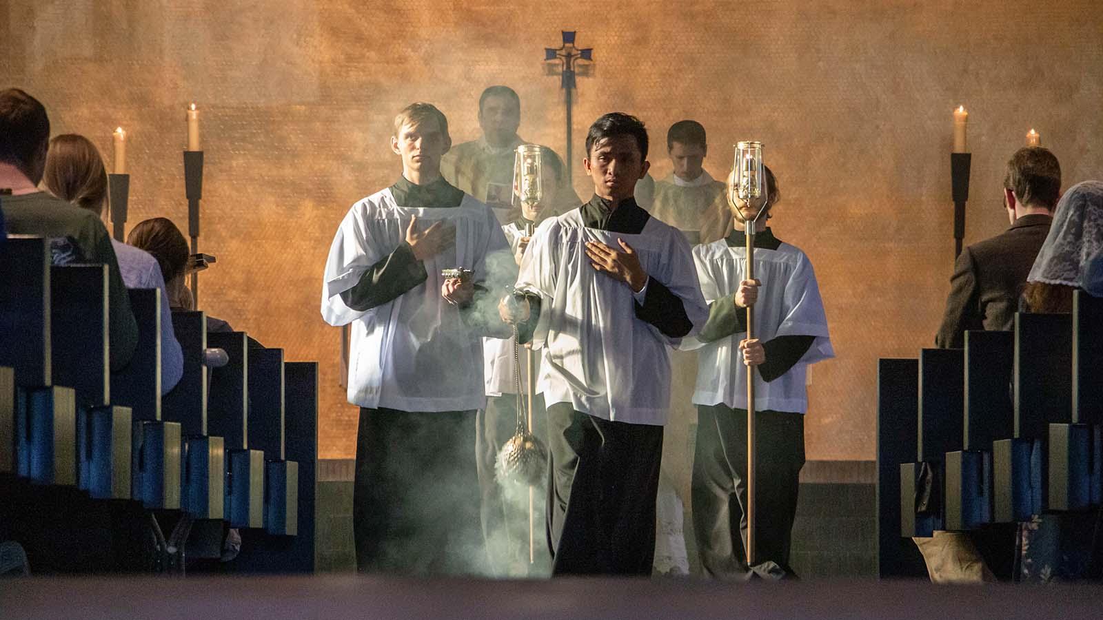 Student carrying a thurible down the center aisle in mass with two students on either side