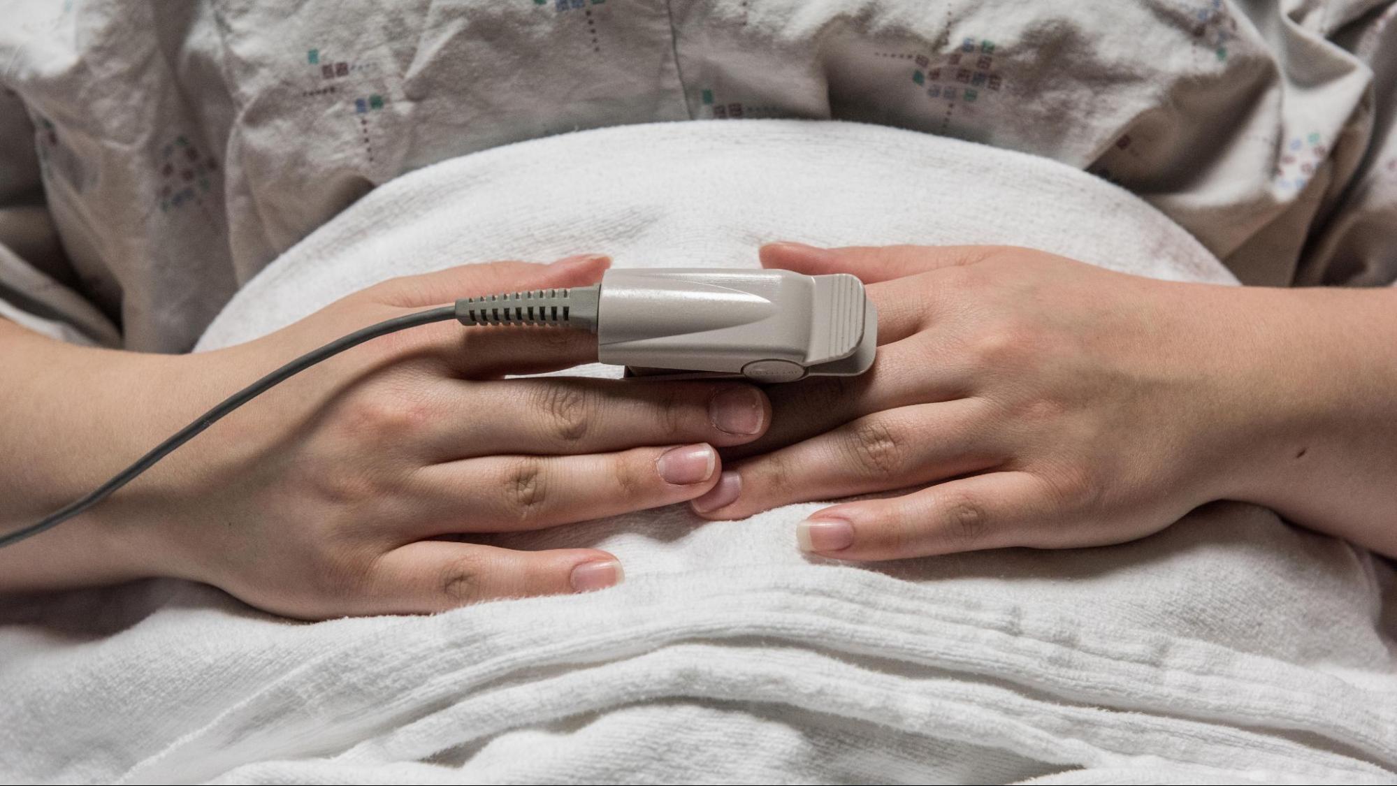 Patient in hospital bed with oxygen monitor attached to their pointer finger.