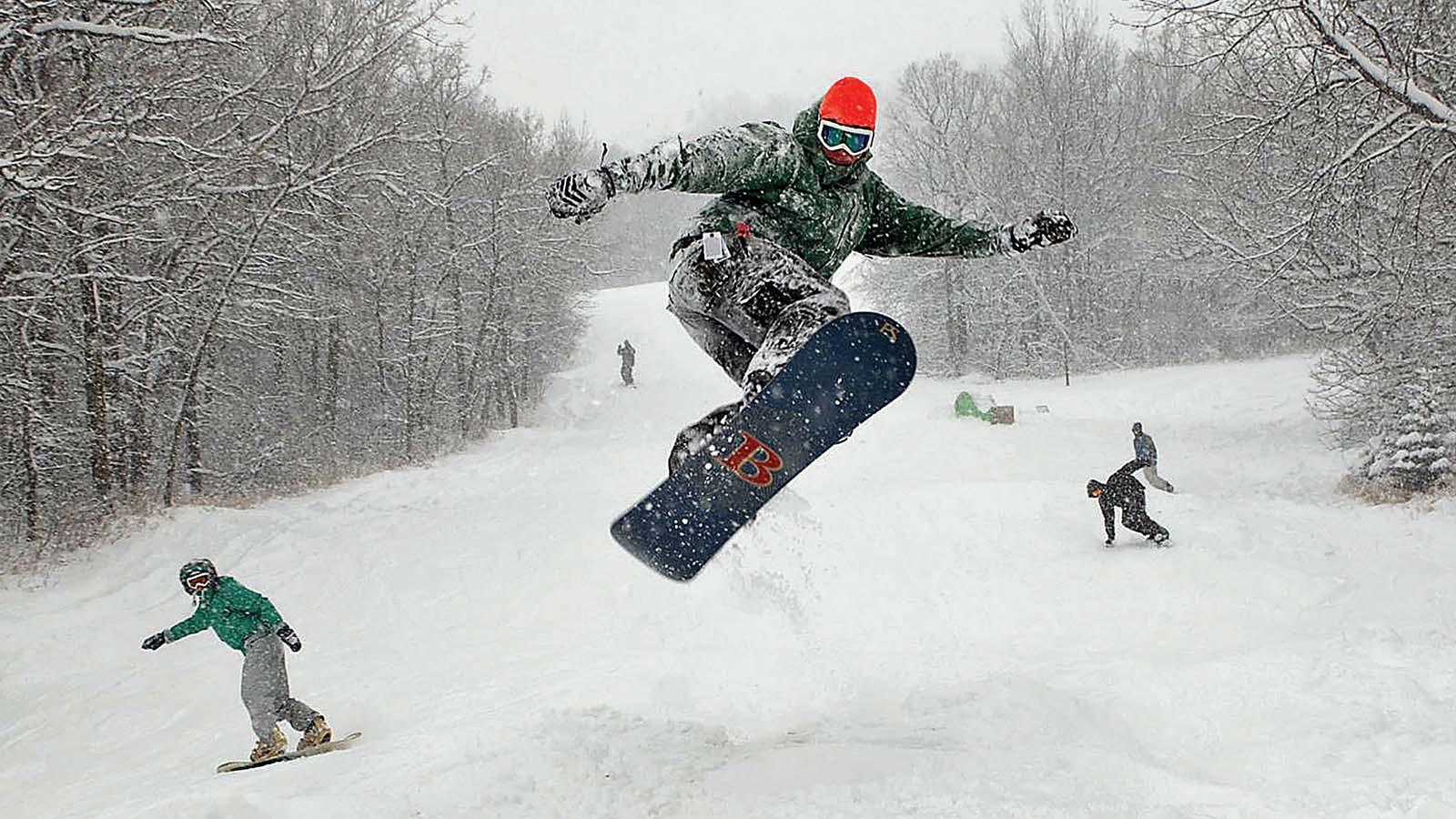 Snowboarder hitting a big jump at Huff Hills with several others snowboarding behind him