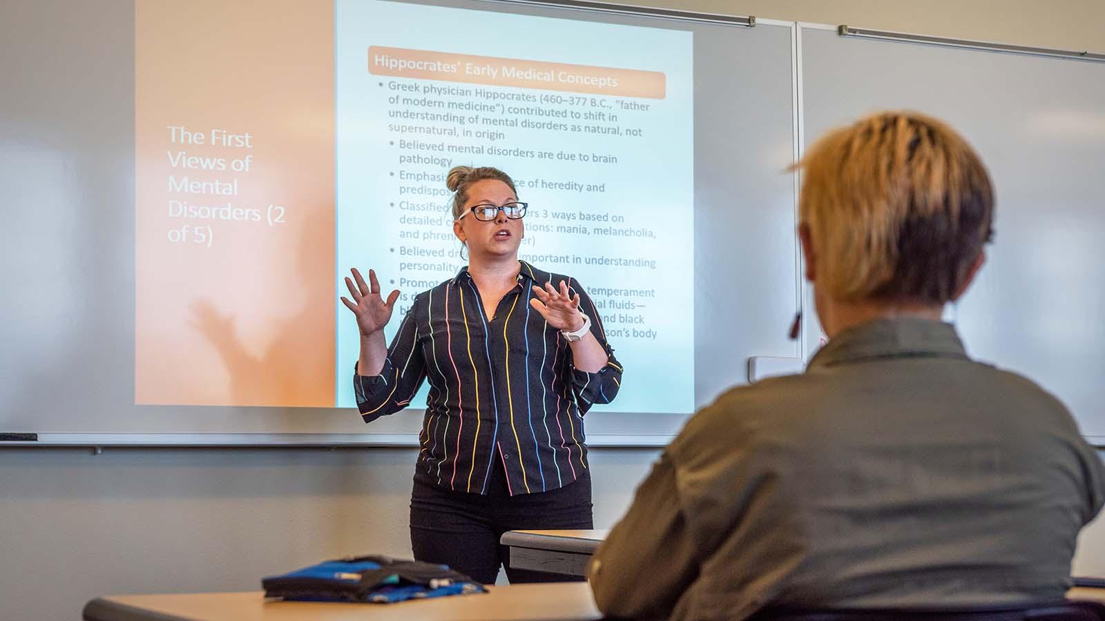 Psychology instructor at the front of class with a mental health disorders presentation behind her