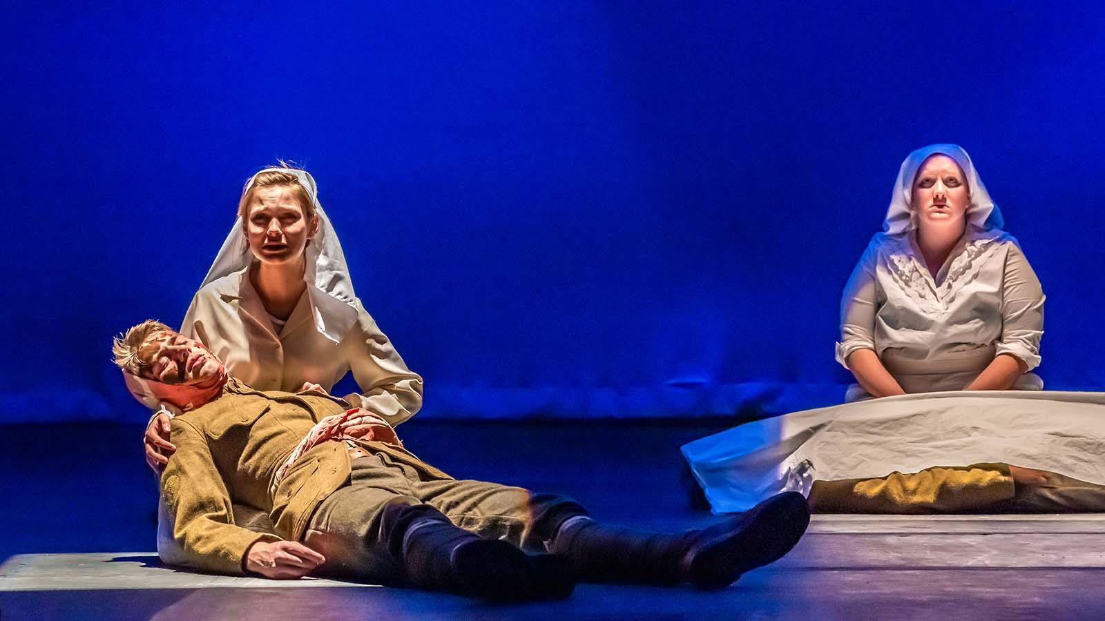 A student actress on stage as a WWI nurse on her knees holding an actor playing a fallen soldier. Another WWI nurse is seated behind them.