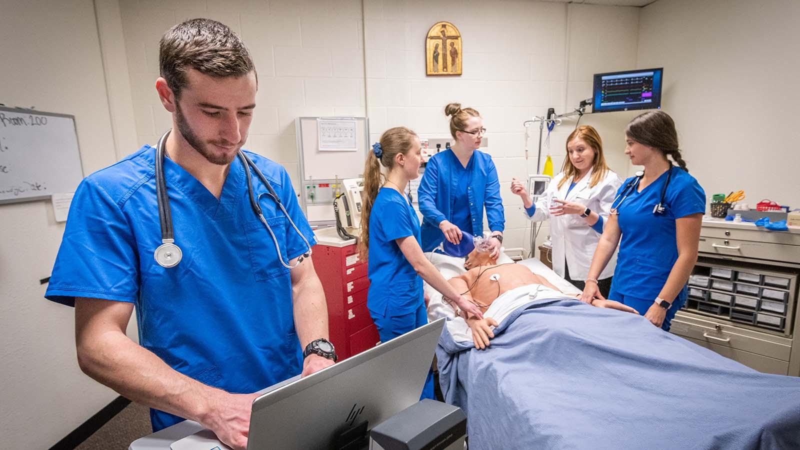 Male nursing student in the foreground on laptop with three nursing students and instructor assessing mannequin in the background