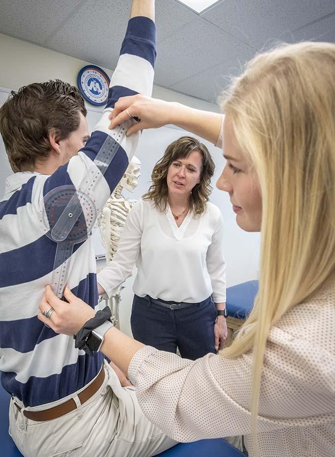 Occupational therapy student assessing shoulder movement of a classmate with the professor looking on