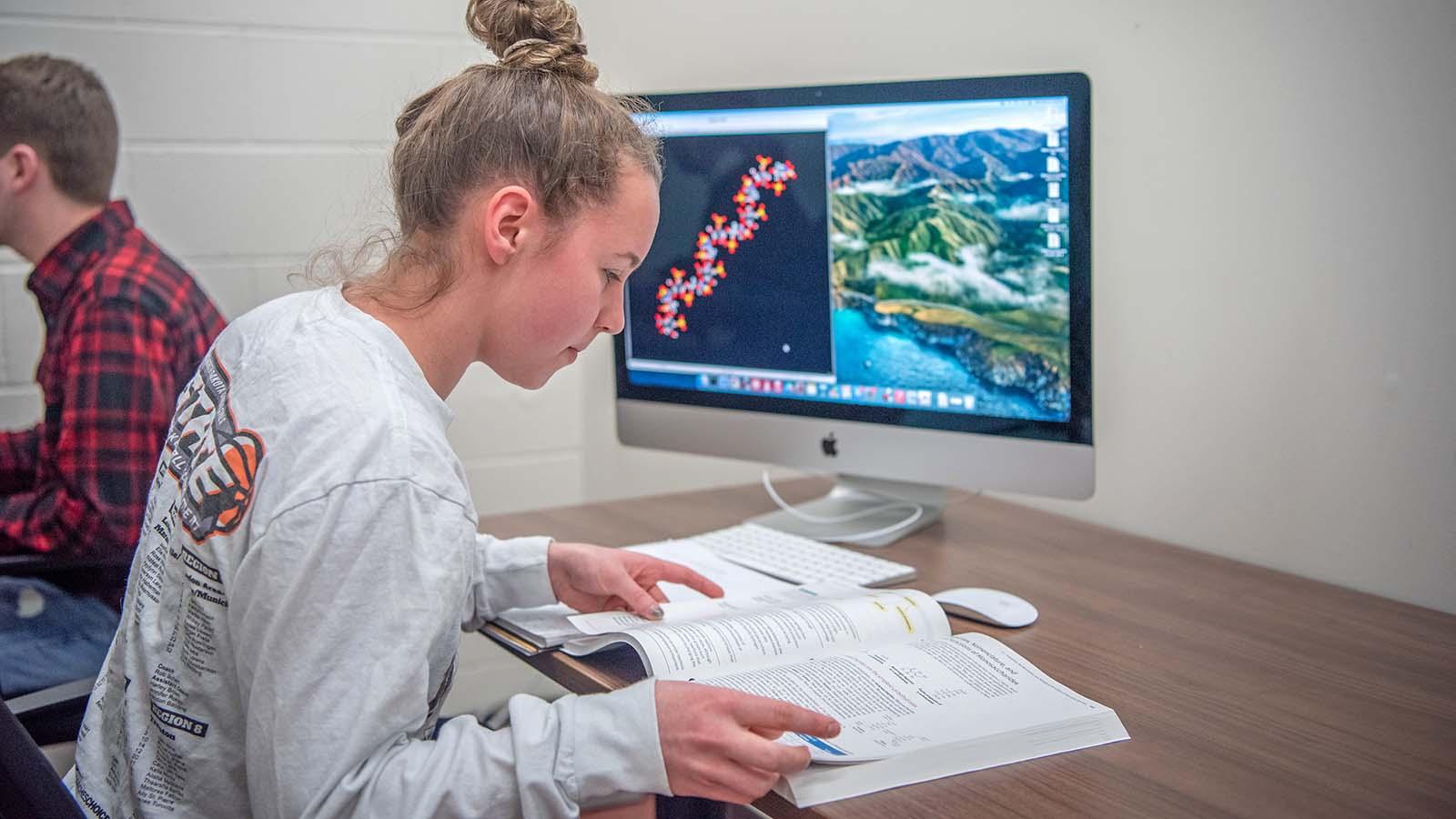 Female student looking at textbook with computer in front of her with DNA strand on the screen.