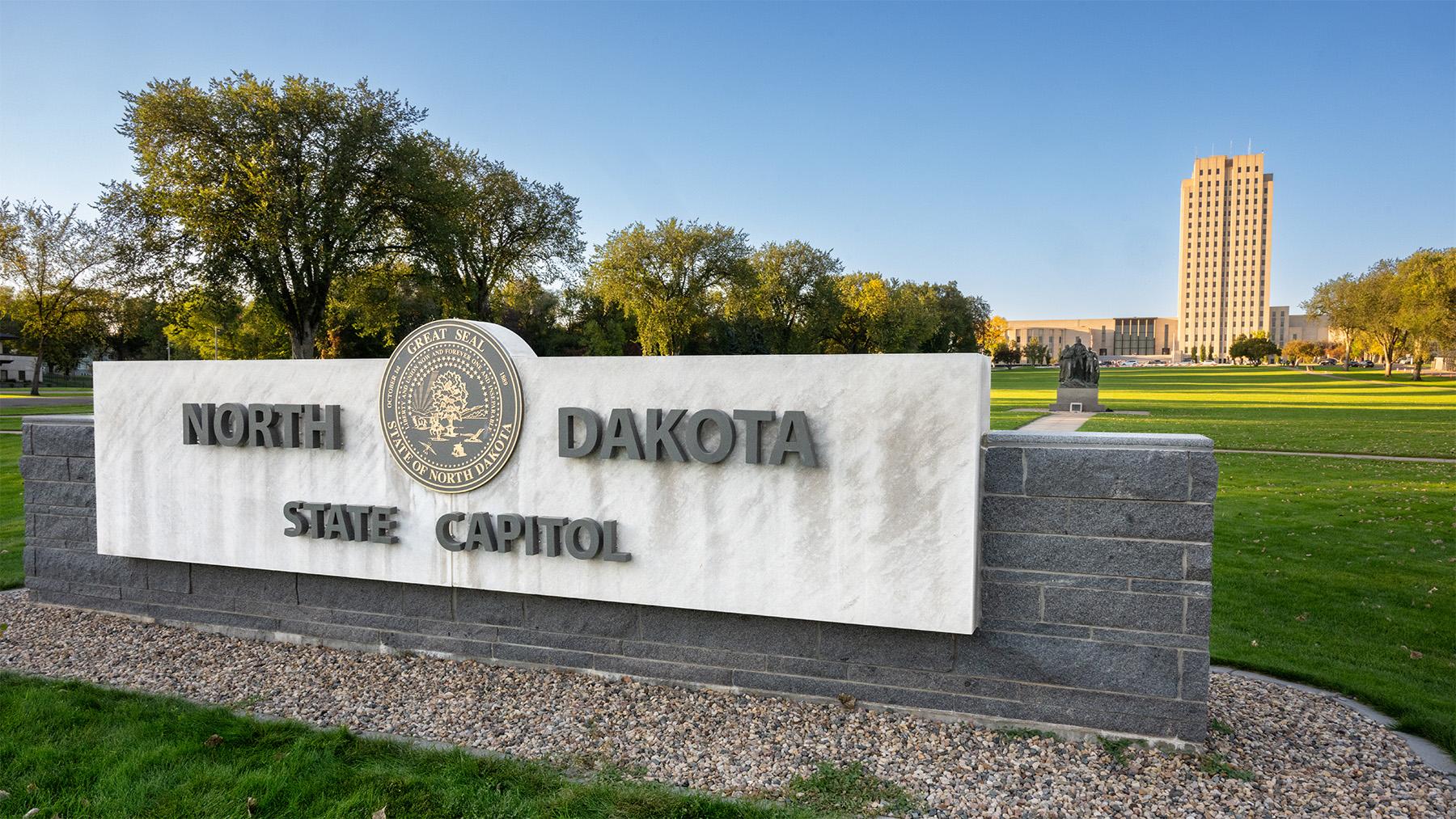 A concrete sign for the ND State Capitol is in forefront with the expansive lawn and capitol building in the background.